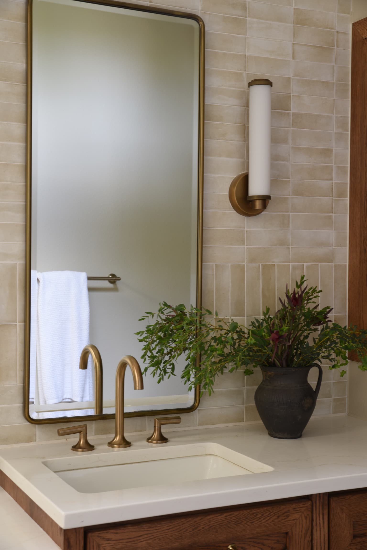 A brass faucet and thin-framed mirror against handmade cream wall tile, with cut greenery in a dark stoneware pitcher.