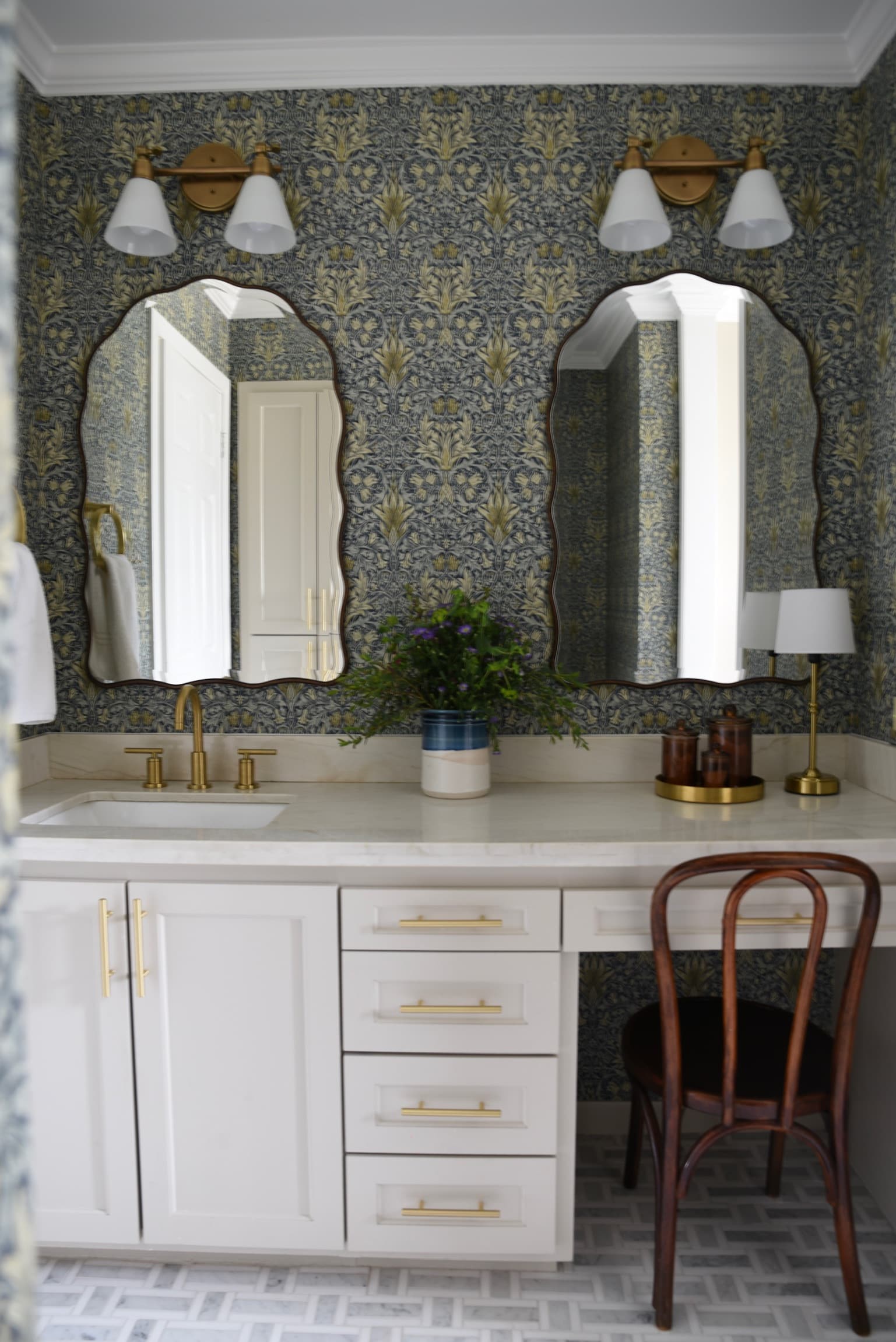 A bathroom papered in a dark botanical print, with twin scalloped mirrors, brass sconces, a white vanity with long brass pulls and a marble herringbone floor.
