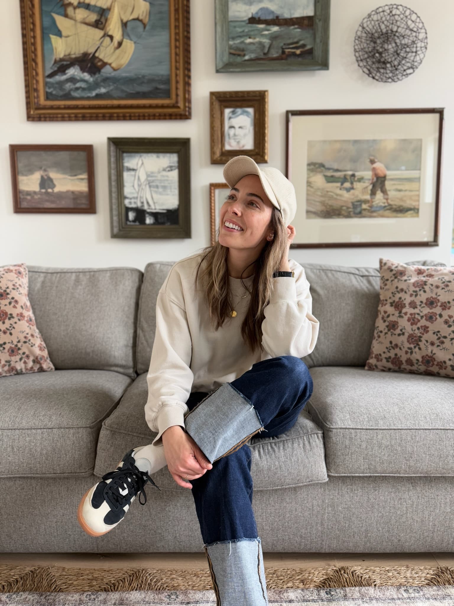 Bekah Madrid seated on a grey sofa in front of a collected gallery wall of framed seascapes and landscapes.