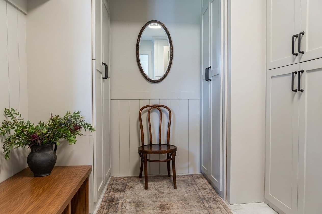 A passage lined in board-and-batten paneling, with a bentwood chair, an oval mirror and a faded vintage runner.