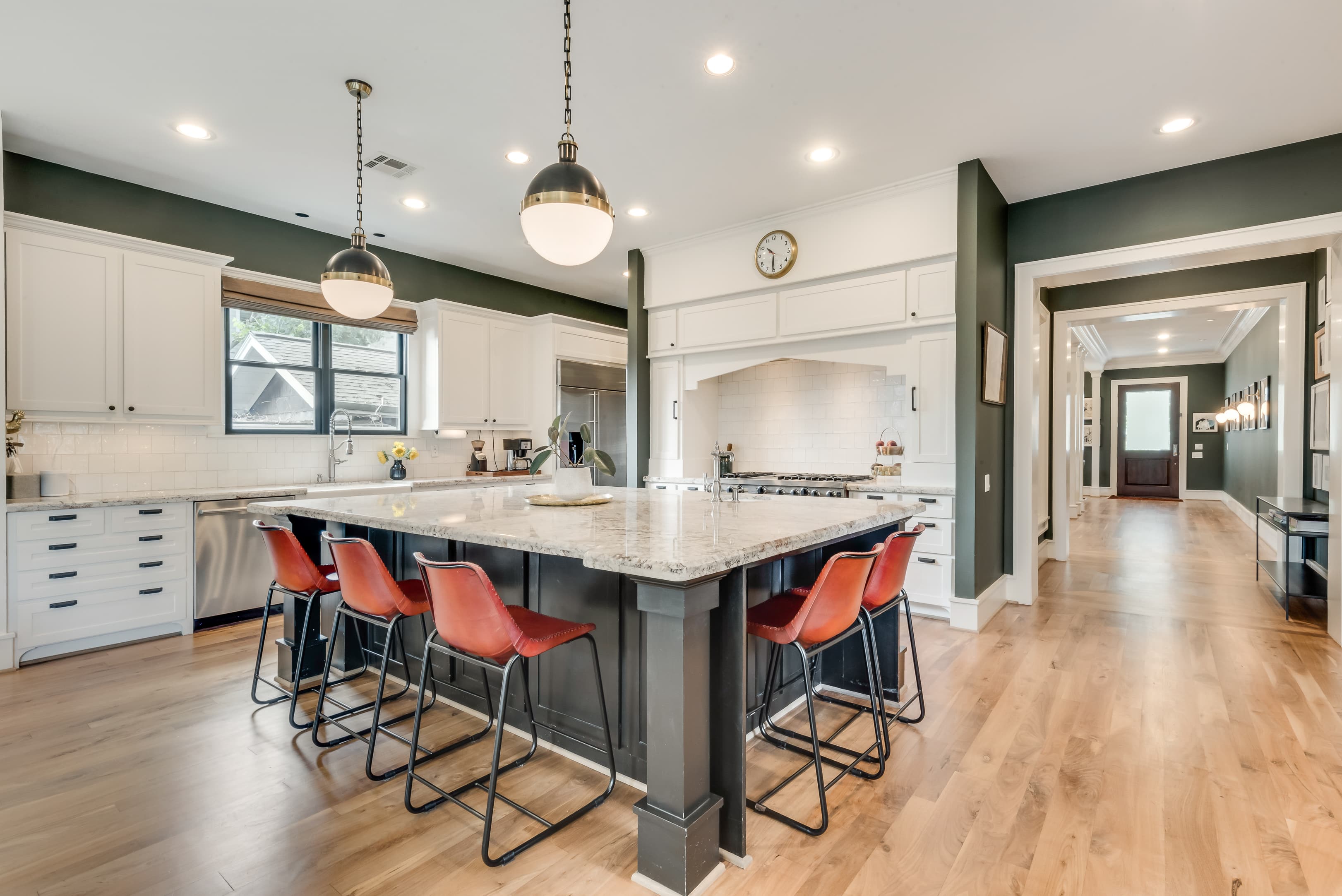 A kitchen in deep olive green with white inset cabinetry and subway tile, a dark island under two brass and milk-glass pendants, and red leather counter stools on pale oak floors.