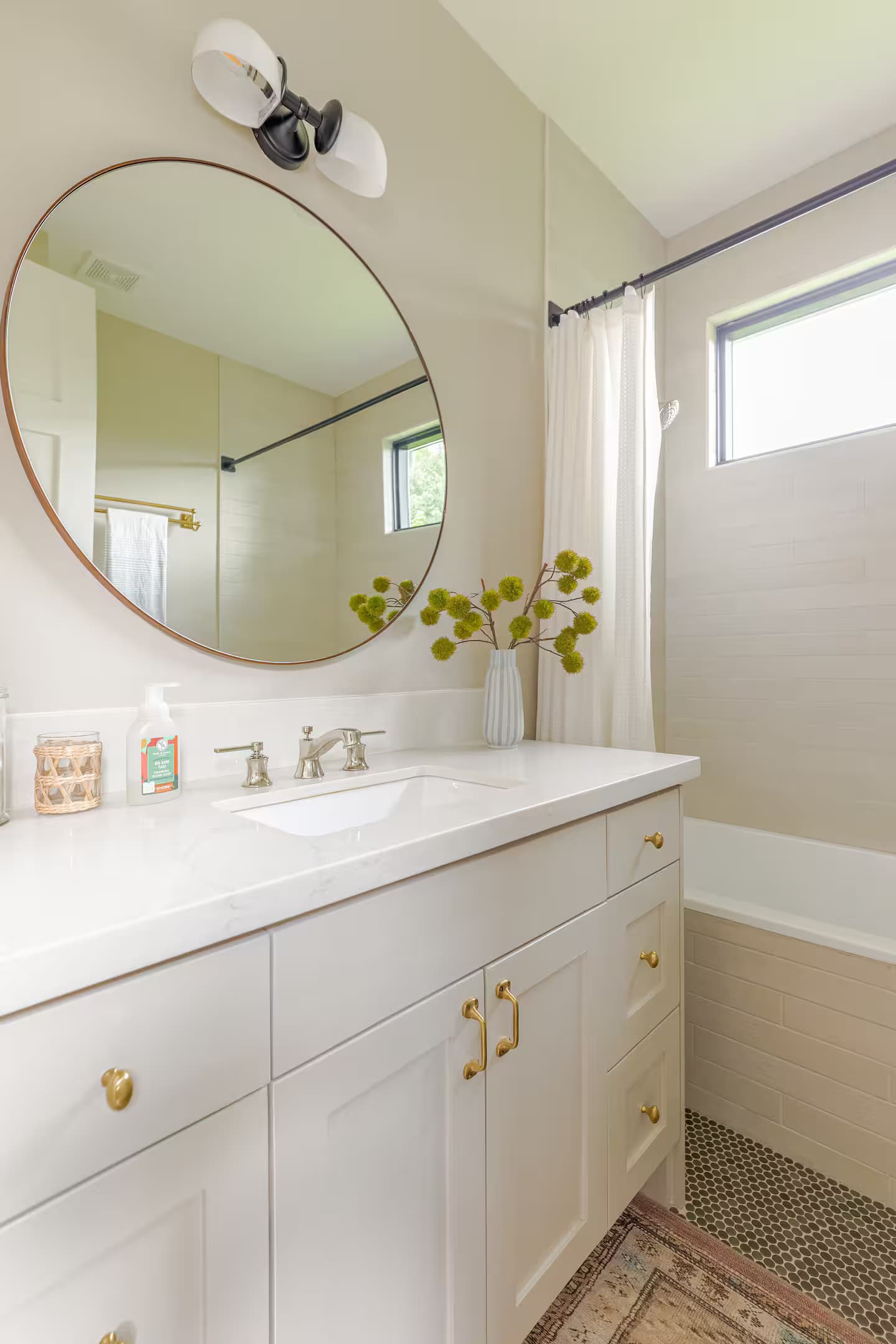A white bathroom with a round metal-framed mirror over a quartz vanity, a fluted vase of green blooms, and a patterned tile floor beside the tub.
