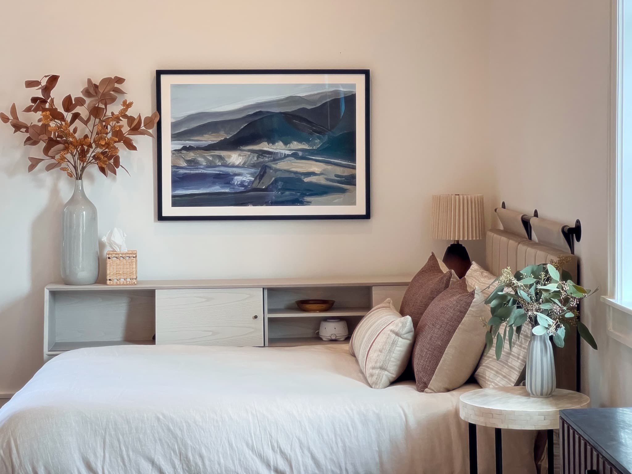 A bed made in white linen below a framed mountain landscape, with dried copper foliage in a tall vase and layered pillows in rust and oatmeal.