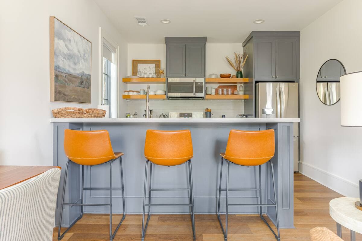 Kitchen with slate blue cabinetry and panelled peninsula, open oak shelving, and three orange leather counter stools.