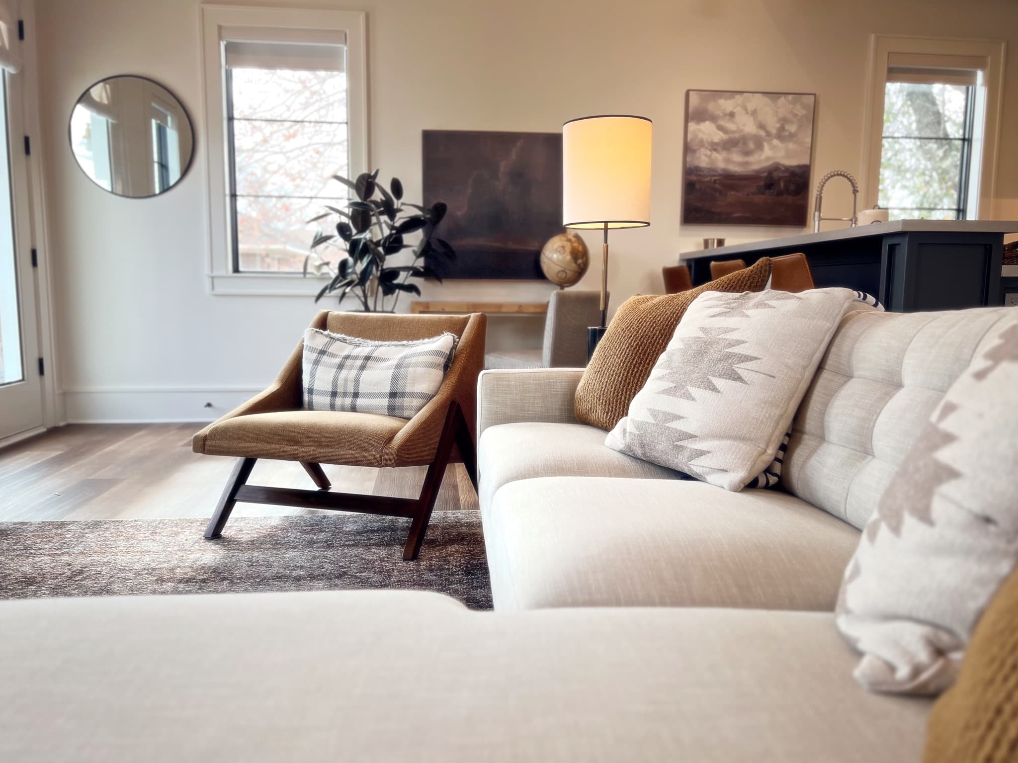 A camel leather lounge chair with a plaid pillow seen past the arm of a pale sofa, with a lit floor lamp and framed landscape paintings beyond.