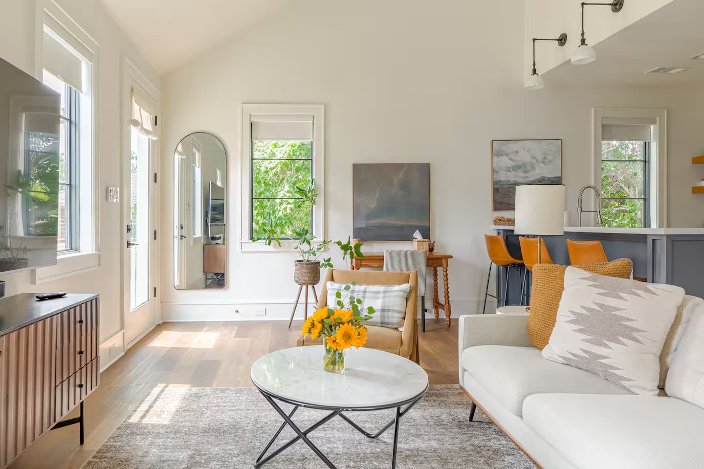 A bright vaulted living room open to the kitchen, with a pale sectional, a round marble coffee table holding sunflowers, an arched floor mirror and orange leather counter stools beyond.