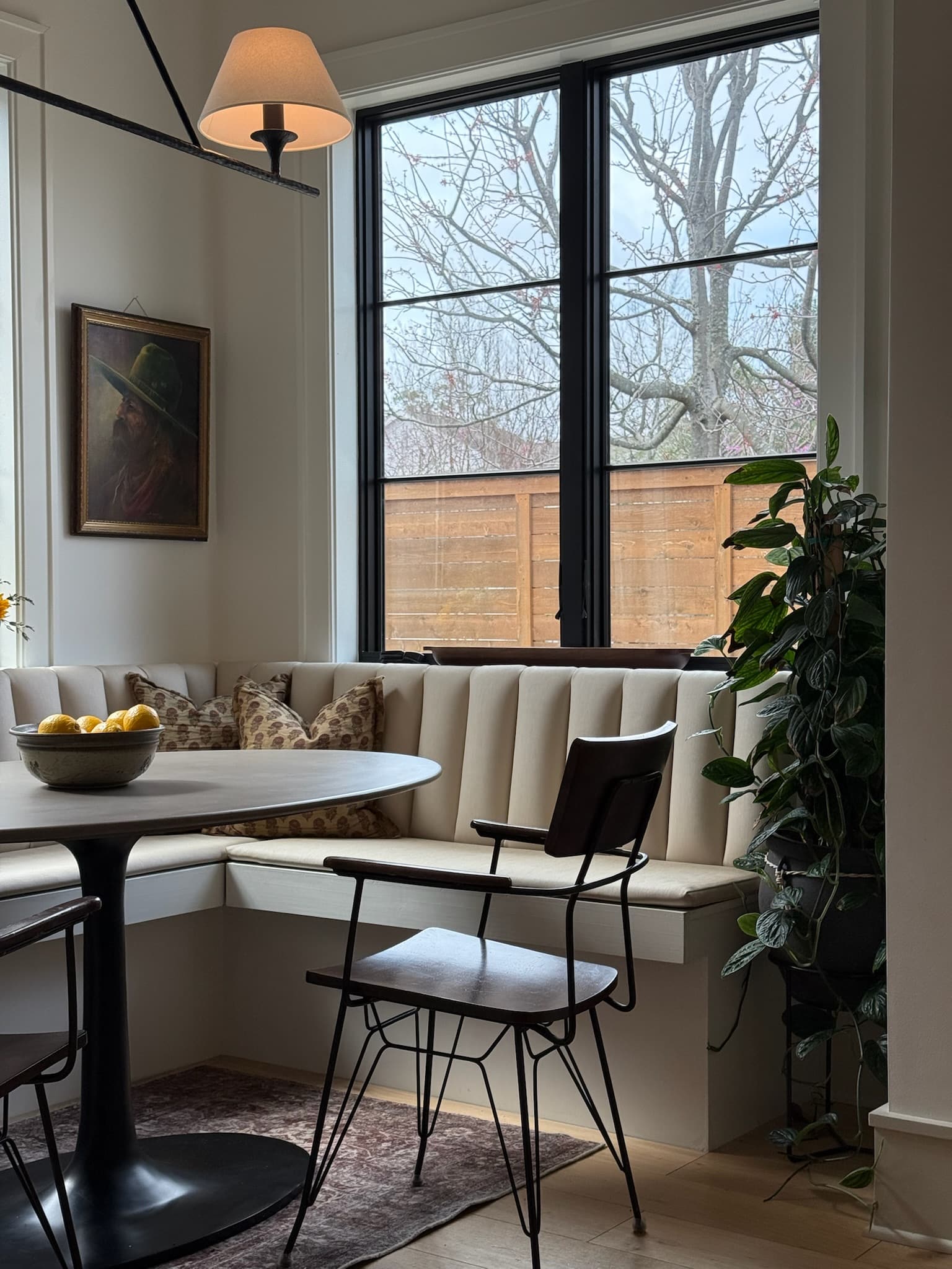 A breakfast nook with a channel-tufted built-in banquette in the corner of two black-framed windows, a round pedestal table and a black wire-back chair.