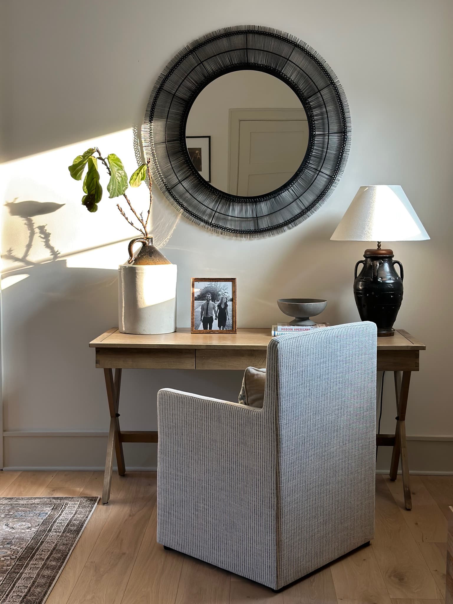An oak writing desk under a large round mirror with a black wire frame, holding a stoneware jug of fig cuttings, a framed photograph and a dark urn lamp, with a striped armchair pulled up to it.