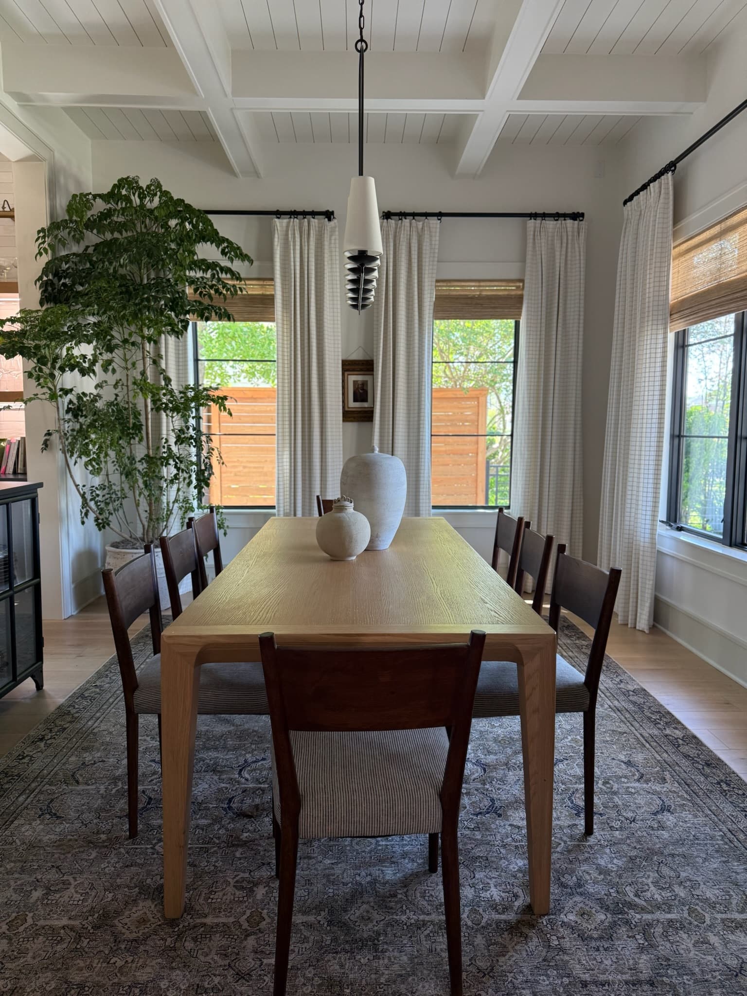 Dining room seen straight on — a long oak table and mixed dining chairs on a faded vintage rug, under a beamed white ceiling, between tall black-framed windows.