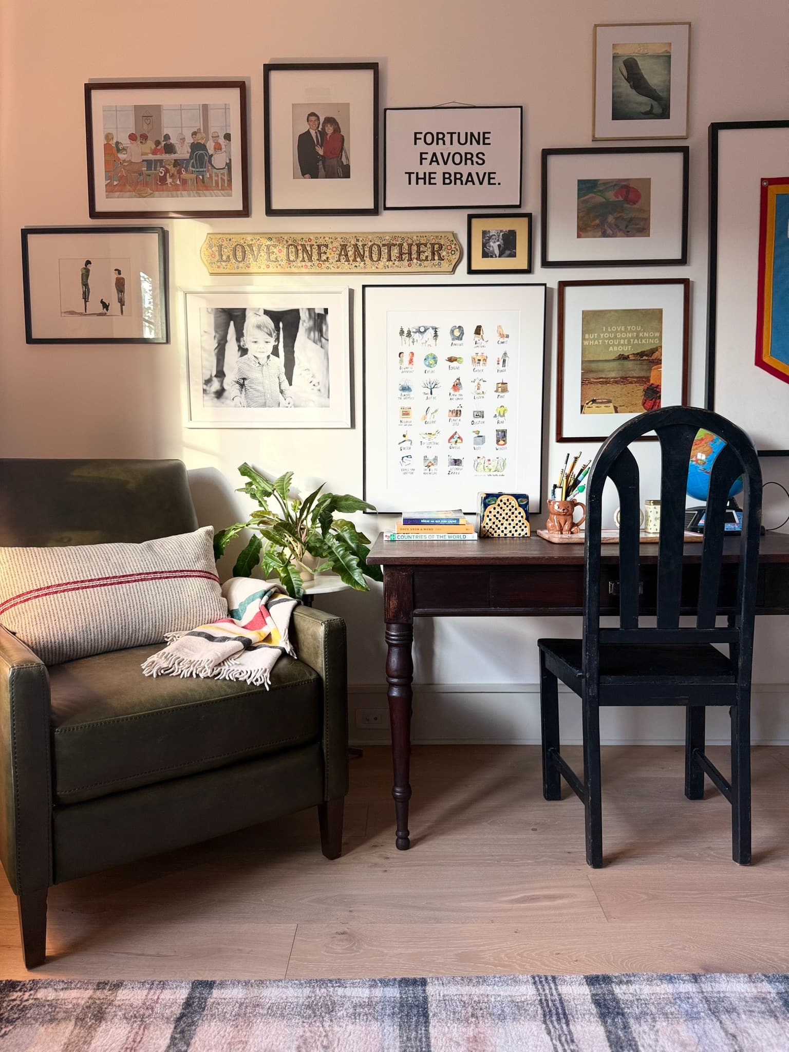 A collected gallery wall of framed drawings, prints and photographs above an antique writing desk, a black spindle chair and an olive green armchair.