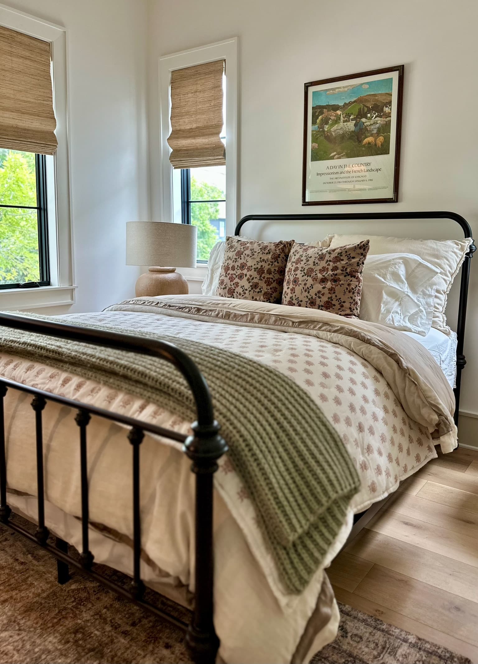 A guest bedroom with a black iron bed made in oatmeal and cream layers with a sage throw, below a framed landscape print.