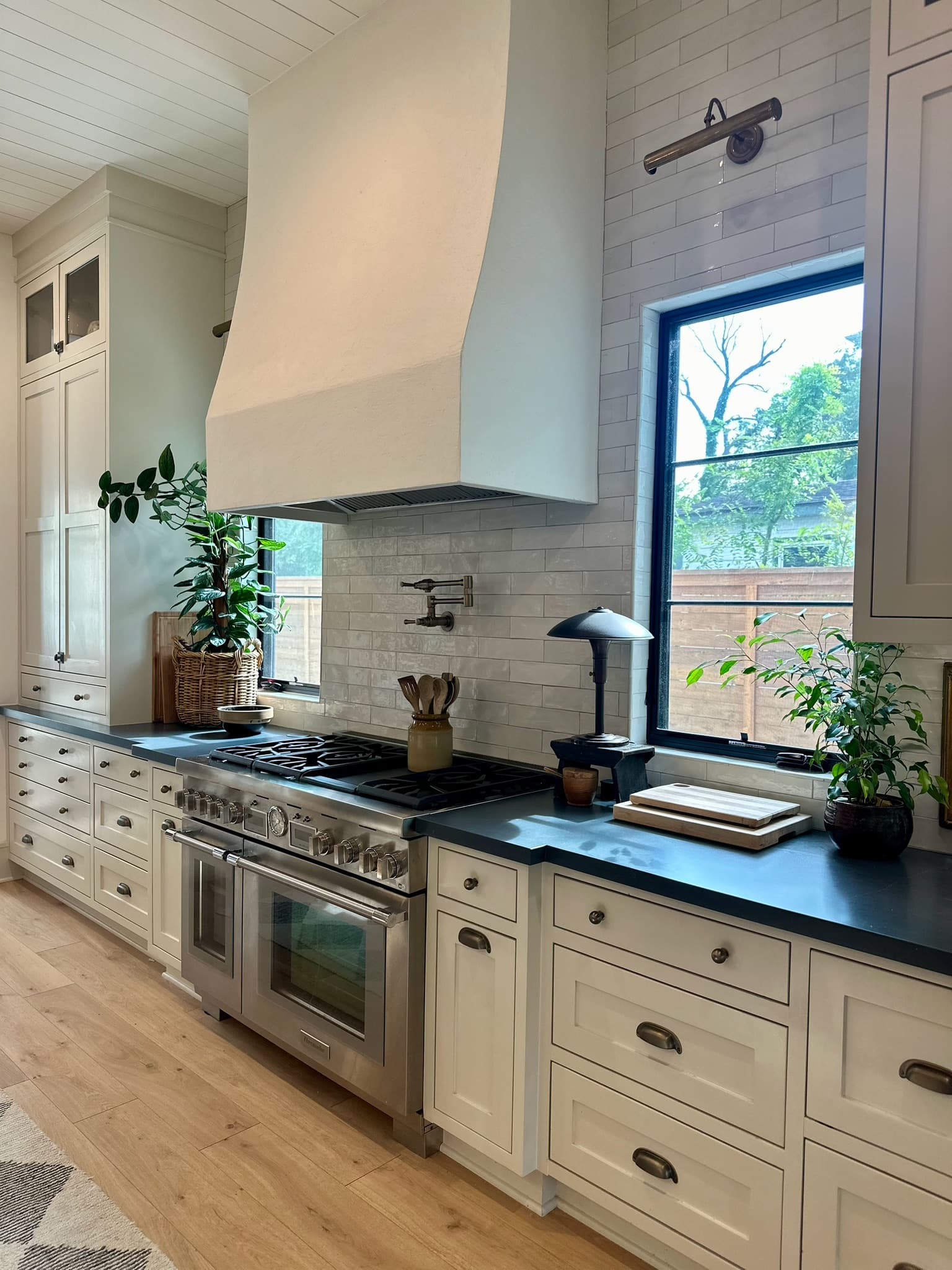 Kitchen range wall with a plaster hood over a stainless range, cream inset cabinetry with dark soapstone counters, and a marble tile backsplash beside a black-framed window.