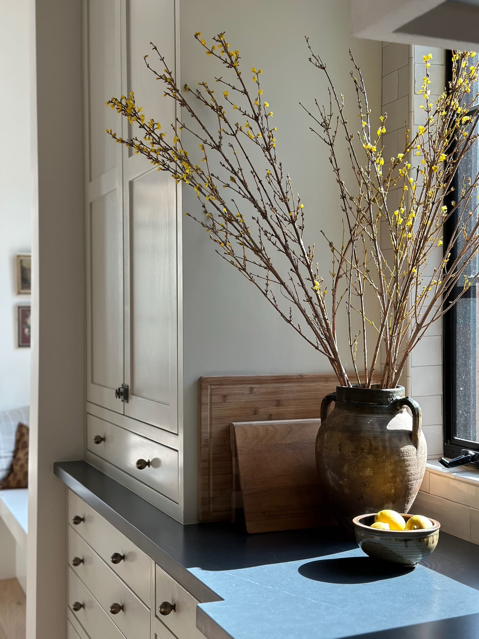 Forsythia branches in an antique olive jar on a dark soapstone counter, beside wooden cutting boards and a bowl of lemons.