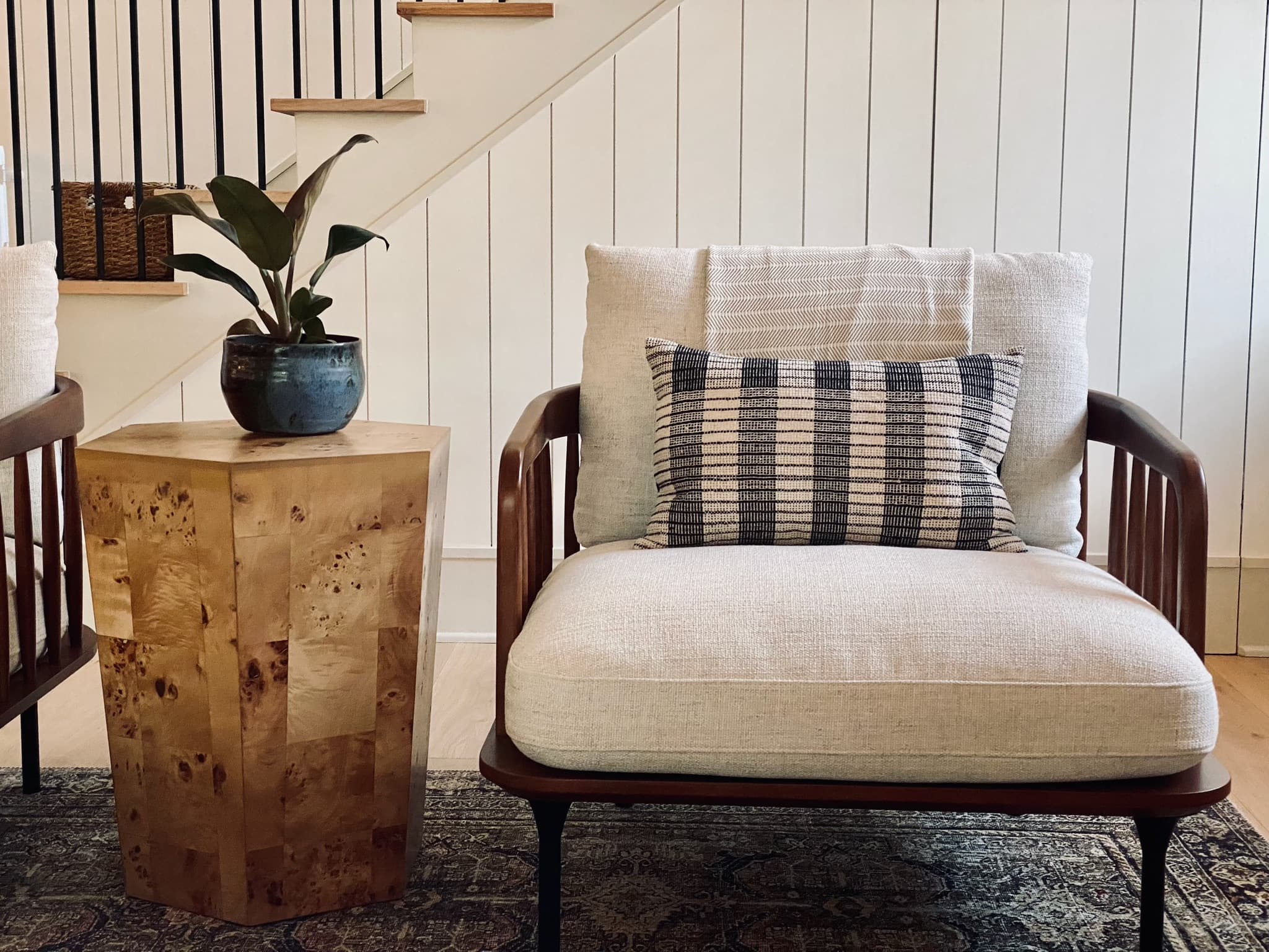 A cane and walnut lounge chair with a striped lumbar pillow beside a burl wood side table, against a white shiplap stair wall.