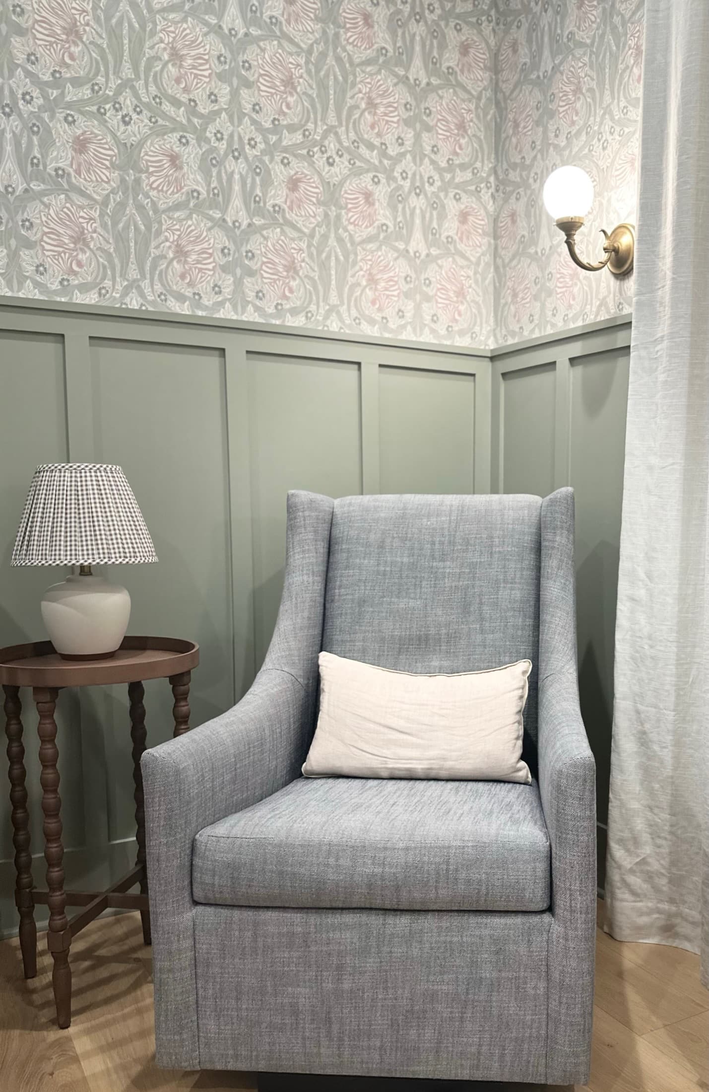A nursery corner with sage green wall paneling below a pale floral wallpaper, a blue-grey slipper chair, a bobbin side table and a gingham lampshade.