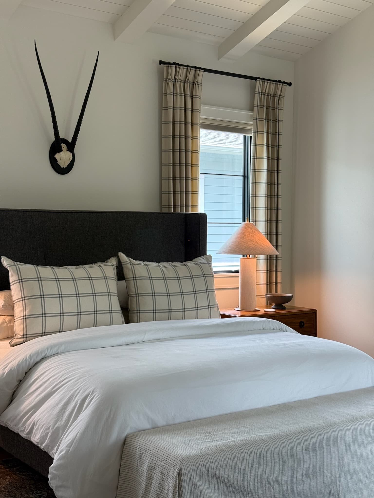 Bedroom with a charcoal upholstered bed and matching windowpane check pillows below a beamed ceiling, beside a window looking onto the garden.