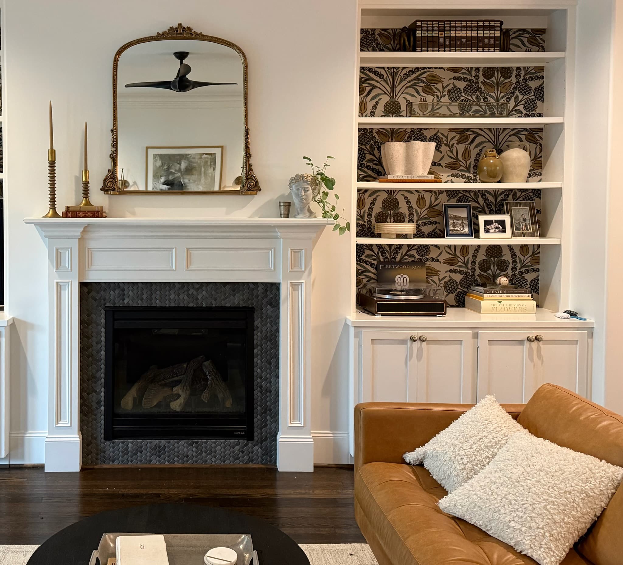 A white panelled fireplace with a dark mosaic surround and gilt mirror, beside built-in shelves papered in a dark botanical print, with a tan leather sofa in front.