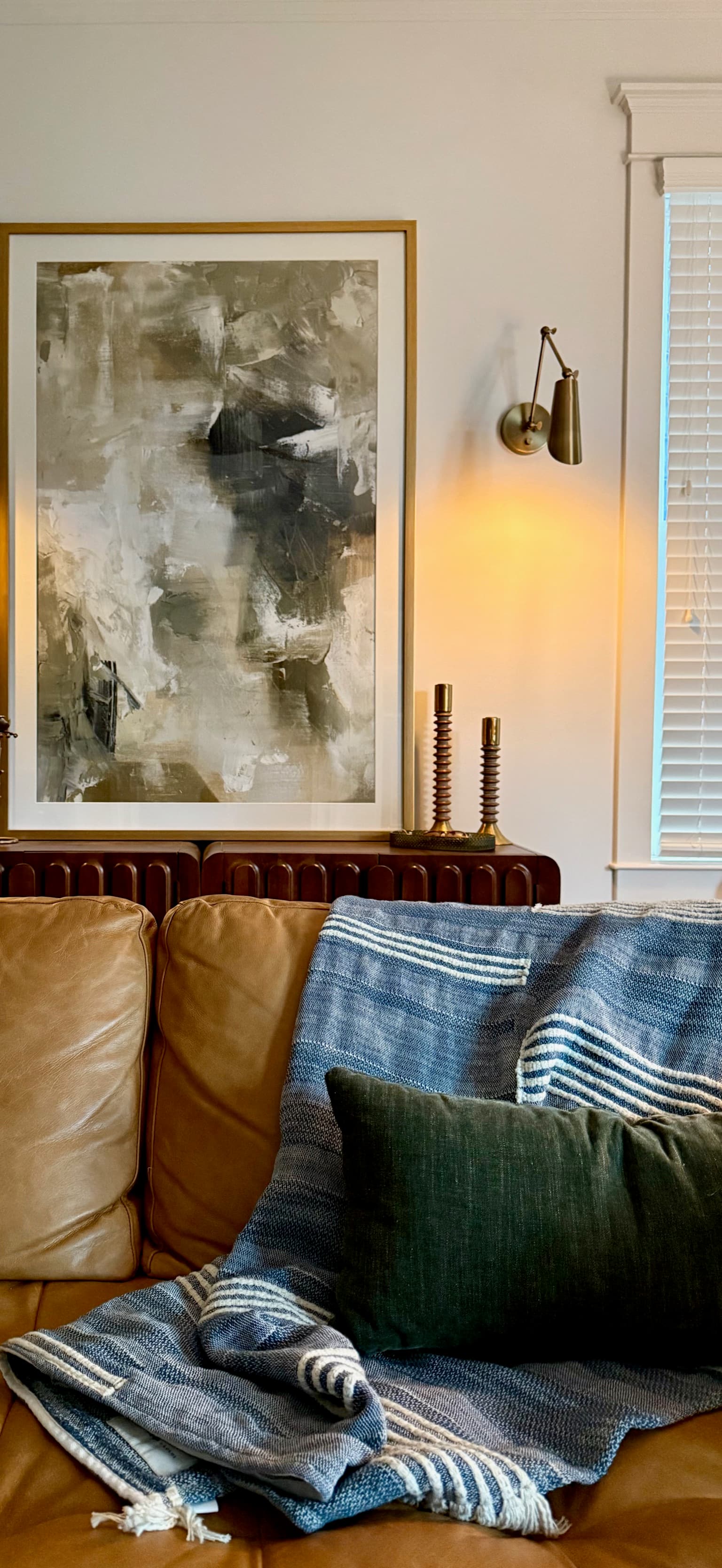A framed abstract painting in oatmeal and charcoal above a fluted walnut console with brass candlesticks, lit by a brass wall sconce, over a camel leather sofa layered with a blue striped throw and a dark green linen pillow.