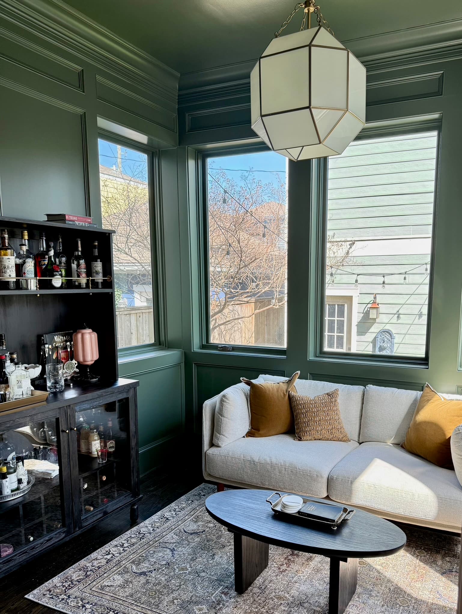 A sunroom painted out in deep olive green — walls, paneling and window frames — with a pale sofa in ochre and oatmeal pillows, a black oval coffee table and a glass lantern pendant.