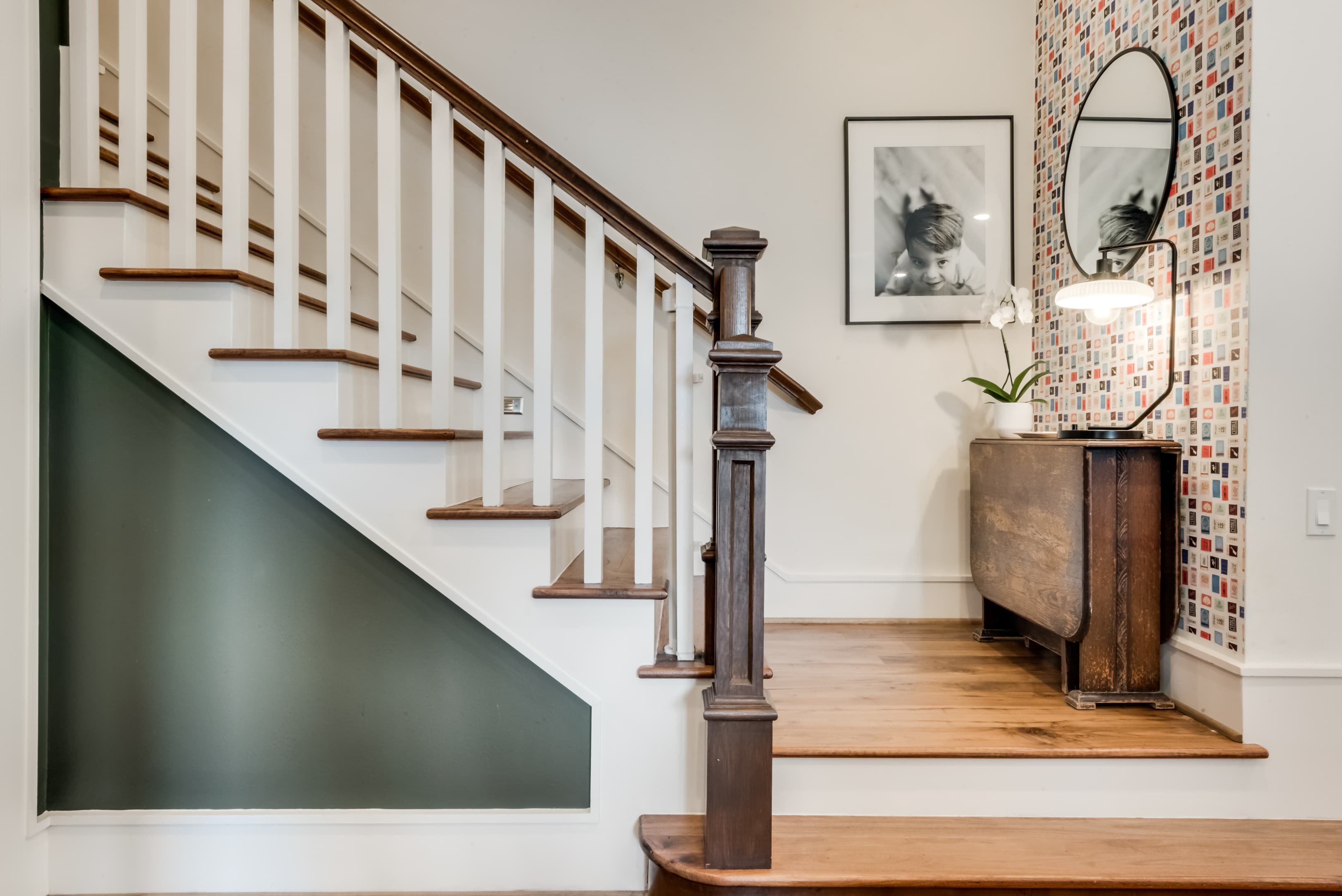 A stair hall with a dark green painted stair skirt, white balusters and a walnut handrail, beside a papered alcove holding an oval mirror, a brass sconce and an antique drop-leaf console.