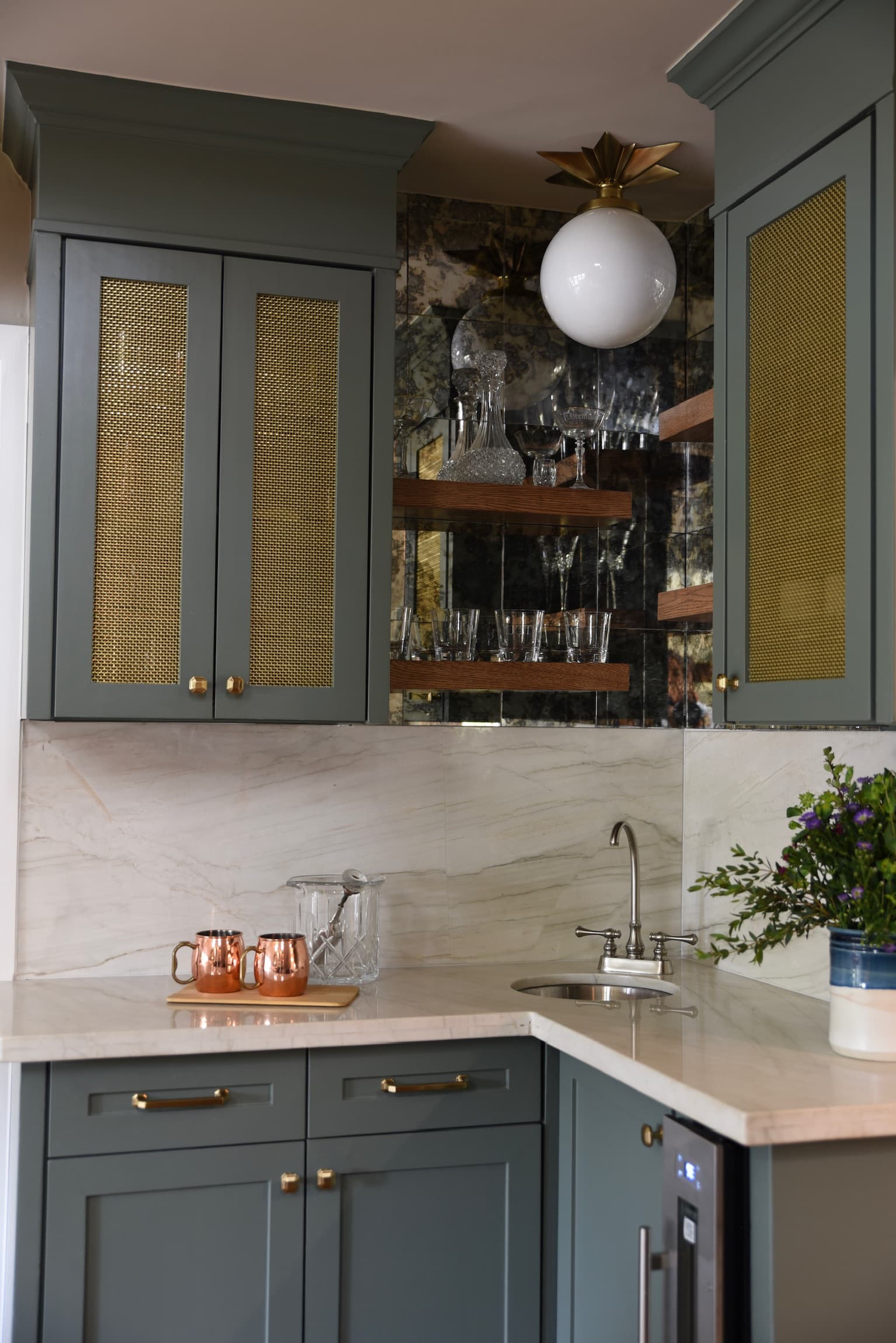 A wet bar in deep teal cabinetry with brass mesh cabinet fronts, walnut floating shelves against an antiqued mirror backsplash, marble counters and a brass and glass ceiling light.